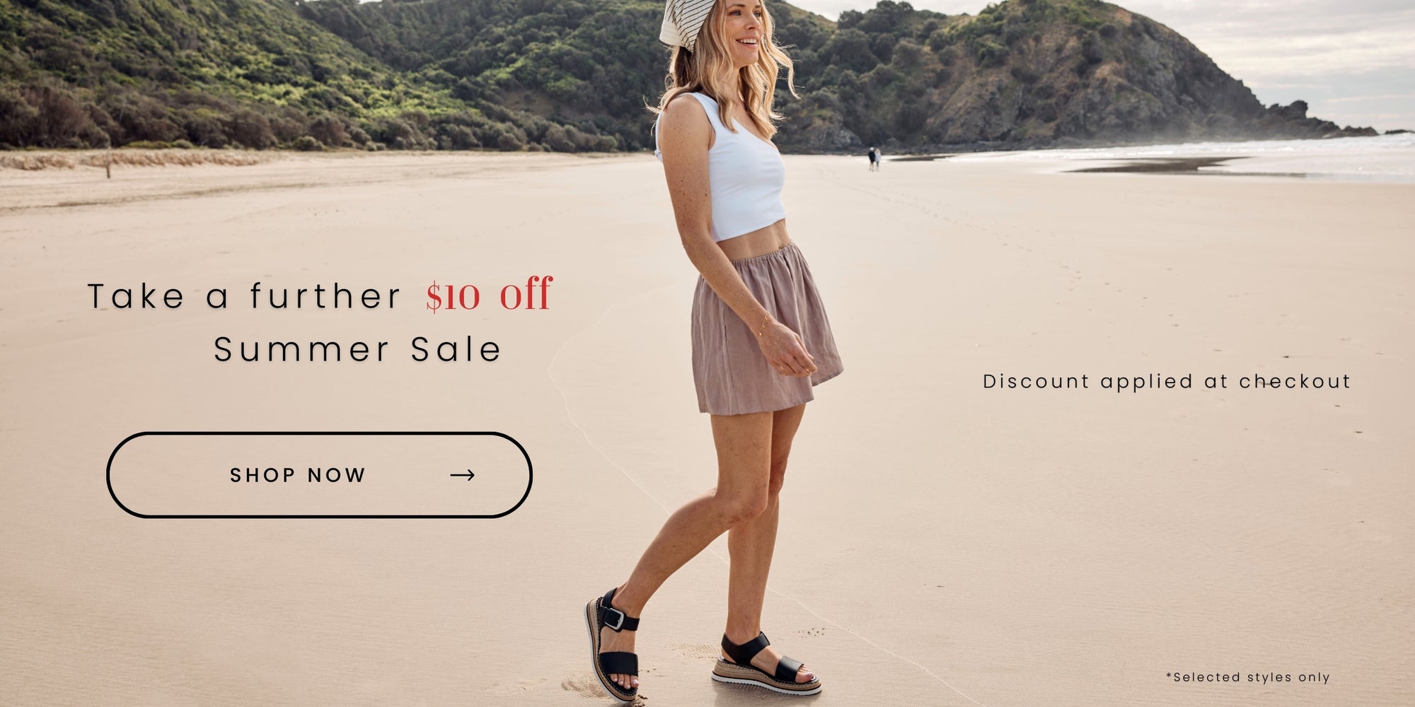 Woman walking on the beach wearing black sandals with a buckle. Text reads "Take a further $10 off Summer Sale. Shop Now. Discount applied at checkout. Selected styles only.'